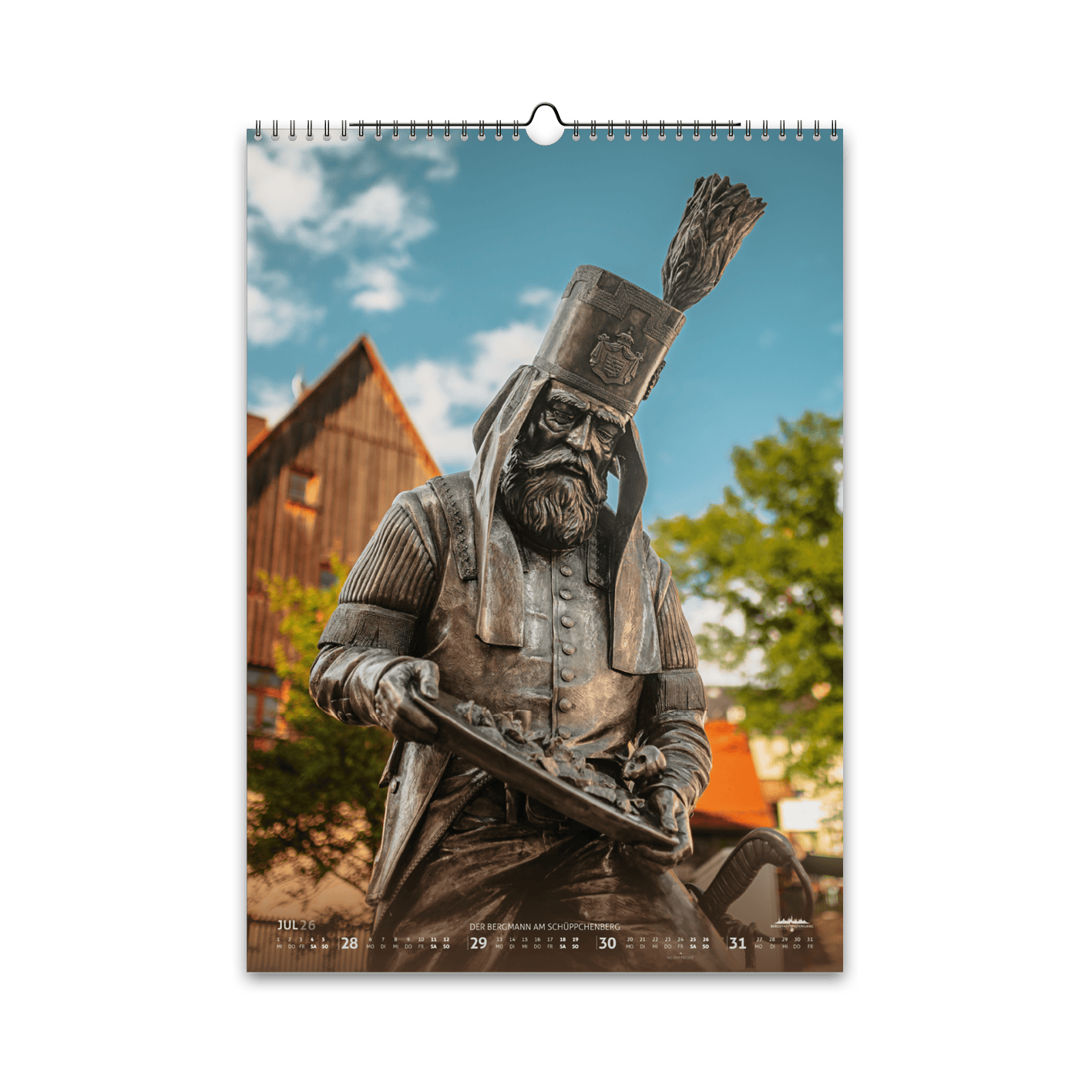 Juli-Motiv des Bergstadtkalenders 2026 im DIN-A3-Hochformat: die detailreiche Bergmann-Figur des Silberwegs am Schüppchenberg in Freiberg, im warmen Sommerlicht vor historischen Gebäuden.