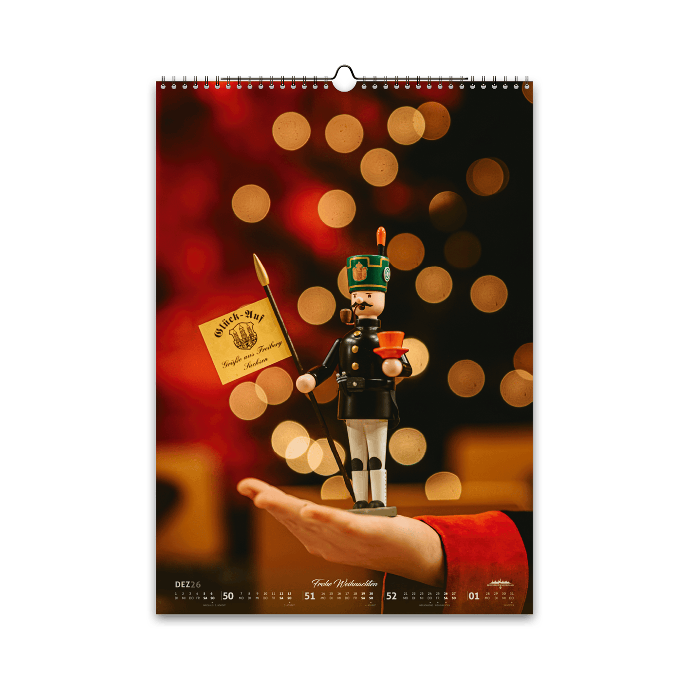 Dezember-Motiv des Bergstadtkalenders 2026 im DIN-A3-Hochformat: eine erzgebirgische Holzfigur des Bergmusikkorps Freiberg, auf einer Hand präsentiert, vor weihnachtlichem Lichterglanz in warmen Gold- und Rottönen.