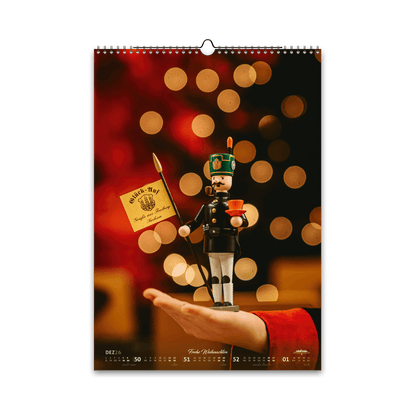 Dezember-Motiv des Bergstadtkalenders 2026 im DIN-A3-Hochformat: eine erzgebirgische Holzfigur des Bergmusikkorps Freiberg, auf einer Hand präsentiert, vor weihnachtlichem Lichterglanz in warmen Gold- und Rottönen.