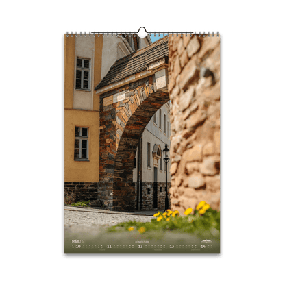 März-Motiv des Bergstadtkalenders 2026 im DIN-A3-Hochformat: das historische Donatstor in Freiberg mit gelb blühenden Frühlingsblumen im Vordergrund und warmem Sonnenlicht auf dem Sandsteinbogen.
