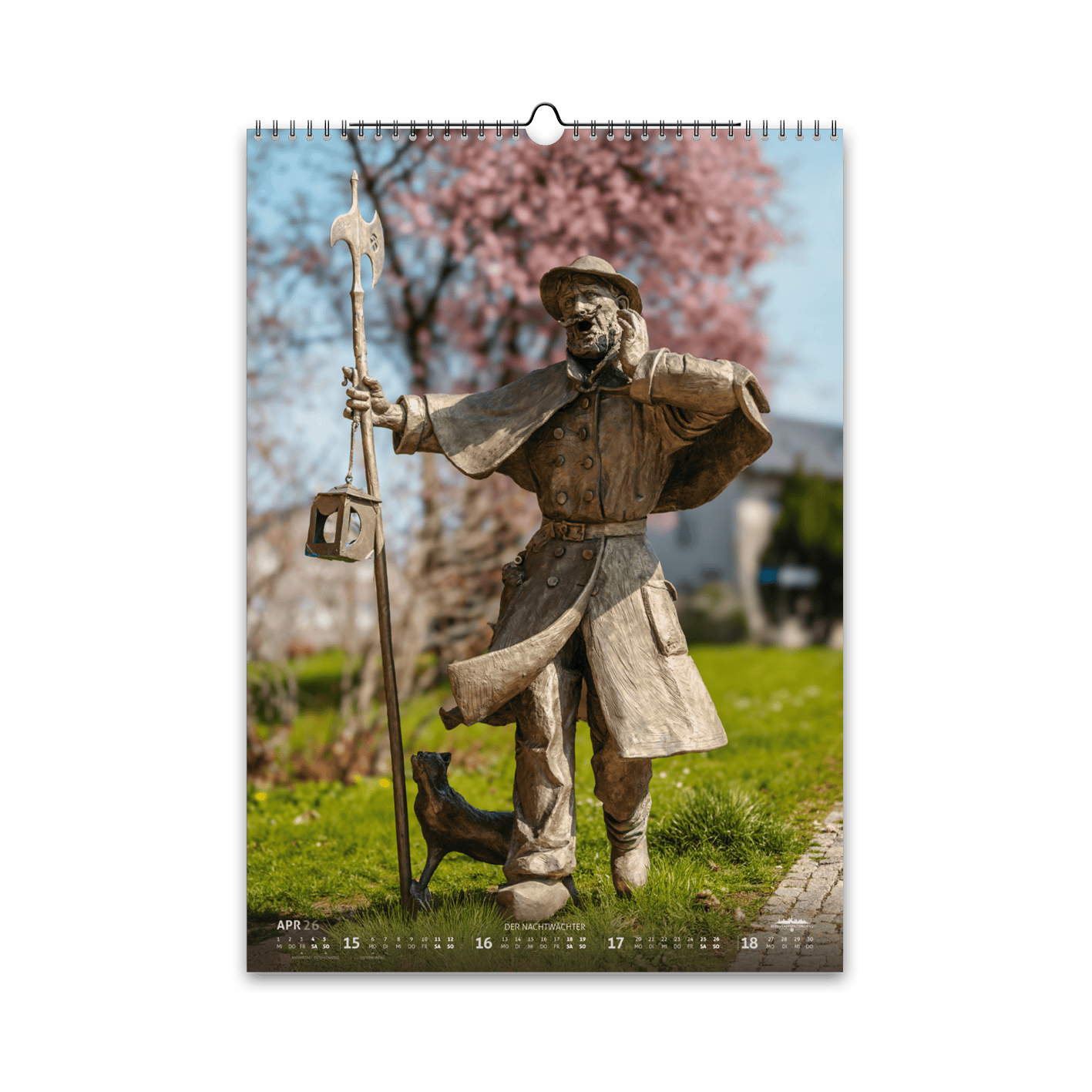 April-Motiv des Bergstadtkalenders 2026 im DIN-A3-Hochformat: die bronzene Nachtwächter-Figur aus der Reihe der Silberweg-Skulpturen in Freiberg, umgeben von blühenden Kirschbäumen im Frühling.