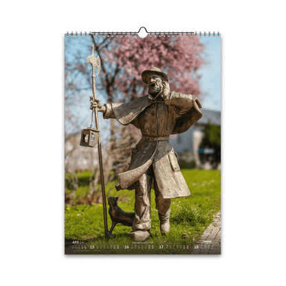 April-Motiv des Bergstadtkalenders 2026 im DIN-A3-Hochformat: die bronzene Nachtwächter-Figur aus der Reihe der Silberweg-Skulpturen in Freiberg, umgeben von blühenden Kirschbäumen im Frühling.