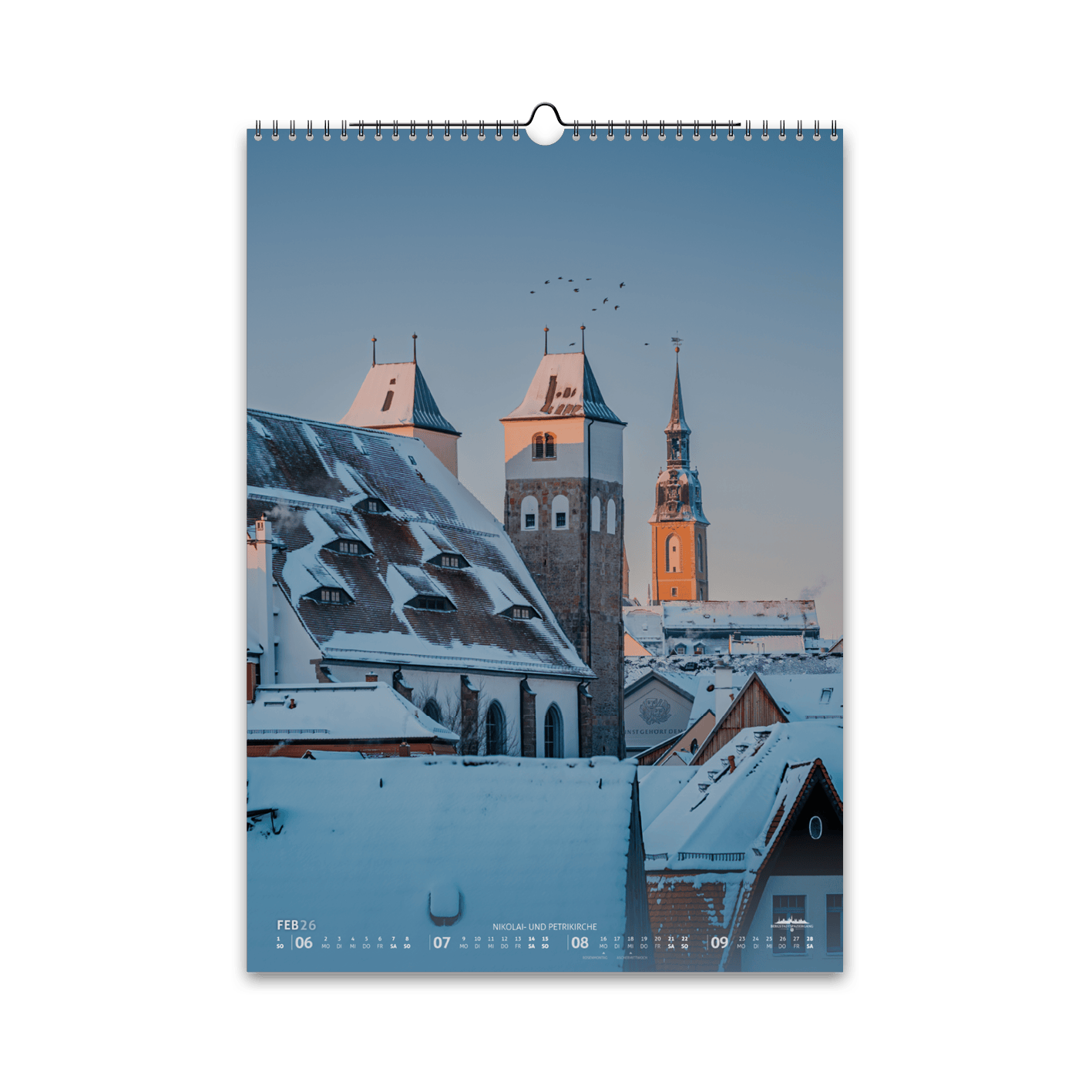Februar-Motiv des Bergstadtkalenders 2026 im DIN-A3-Hochformat: verschneite Dächer von Freiberg mit der Nikolaikirche im Vordergrund und der Petrikirche im Hintergrund, im warmen Licht eines Wintermorgens.