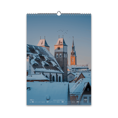 Februar-Motiv des Bergstadtkalenders 2026 im DIN-A3-Hochformat: verschneite Dächer von Freiberg mit der Nikolaikirche im Vordergrund und der Petrikirche im Hintergrund, im warmen Licht eines Wintermorgens.