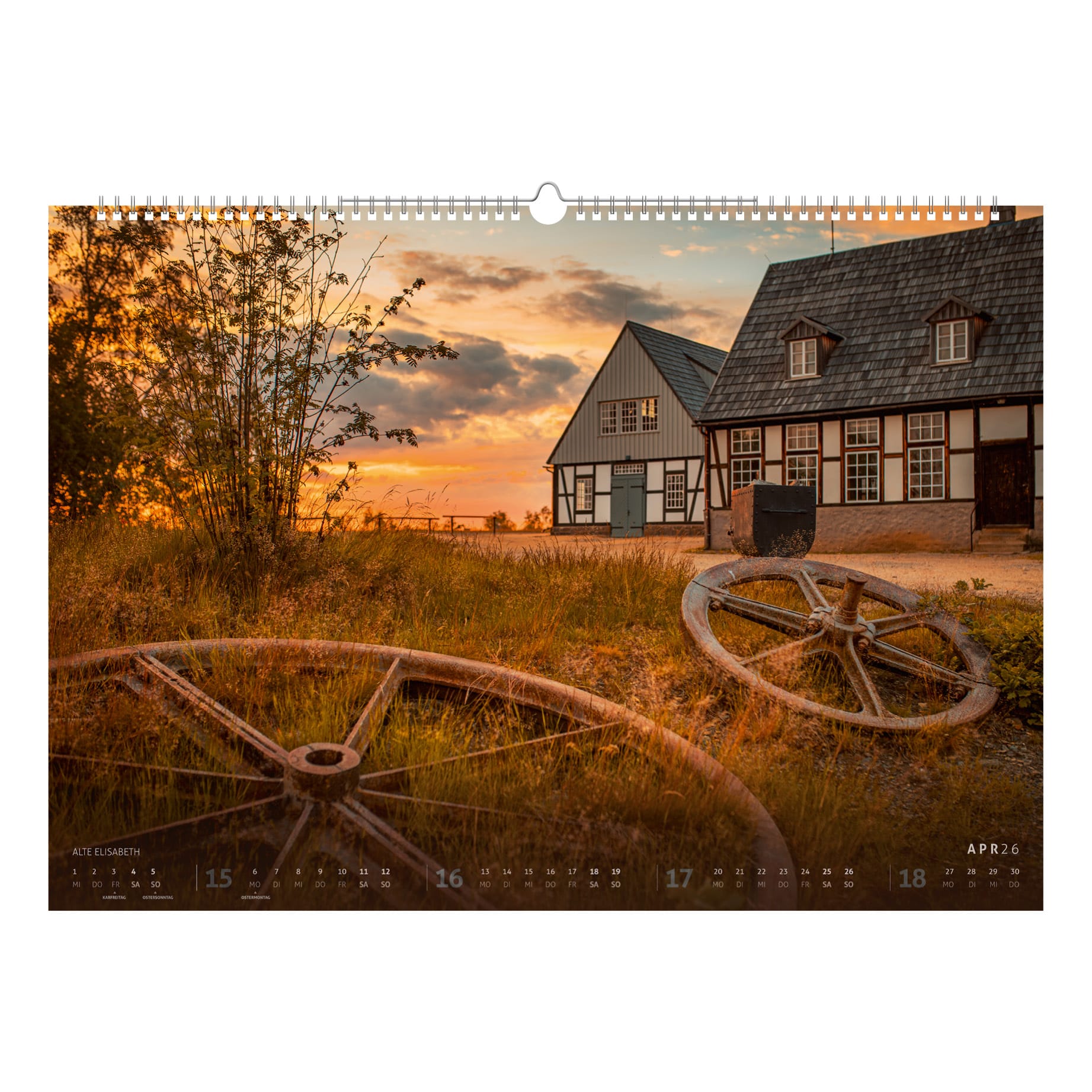 Die historische Förderanlage Alte Elisabeth bei Freiberg im goldenen Abendlicht. Im Vordergrund liegen große Seilscheiben als Erinnerung an die Bergbaugeschichte.