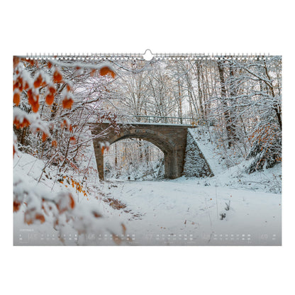 Verschneite Brücke im Freiberger Stadtwald, umgeben von ruhigem Winterwald und zarten orangefarbenen Blättern – Motiv aus dem Bergstadtkalender 2026.